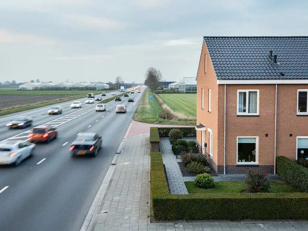 Nieuwgebouwde Nederlandse rijtjeswoning aan drukke meerbaansweg in Westlands polderlandschap, met bakstenen gevel en verzorgde voortuin.