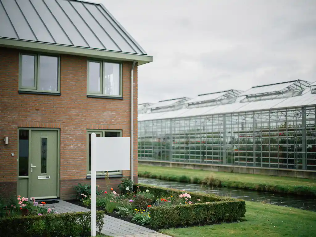 Nieuwbouwwoning te koop in Dutch poldergebied naast glazen kassencomplex, met verzorgde voortuin en overcast daglicht.
